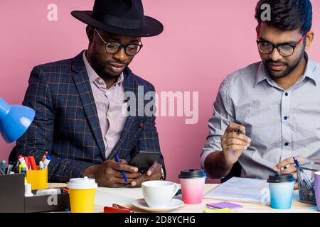 afroamerikanischer Geschäftsmann und indischer Unternehmer diskutieren Geschäftspläne, sitzen am Tisch im Büro, tragen formale Kleidung, lächeln und lachen. Stockfoto