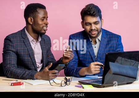 afroamerikanischer junger Geschäftsmann in Anzug zeigt Präsentation seines Projekts auf Laptop an indischen Geschäftspartner, sitzen am Tisch im Büro, holdi Stockfoto