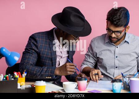 afroamerikanischer Geschäftsmann und indischer Unternehmer diskutieren Geschäftspläne, sitzen am Tisch im Büro, tragen formale Kleidung, lächeln und lachen. Stockfoto