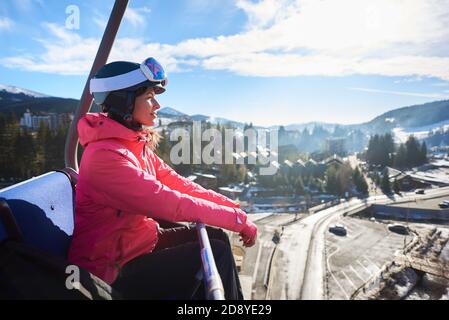 Junge attraktive Frau, Tourist Skifahrer in Helm, Brille und warme Kleidung Reiten in Seilbahn hoch in der Luft auf dem Hintergrund des Skigebiets. Aktiver Lifestyle, Winterurlaub, Sport- und Erholungskonzept. Stockfoto