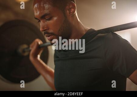 Entschlossener Mann, der im Fitnessstudio die Langhantel auf der Schulter hält. Männliche Bodybuilder arbeiten mit schweren Gewichten in Cross-Training Turnhalle. Stockfoto