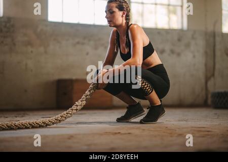 Fitness-Frau sitzt auf dem Boden mit Kampfseil. Frau, die sich nach dem Training im Fitnessstudio in einem alten Lagerhaus ausruht. Stockfoto