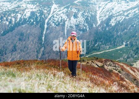 Gekleidet hell orange Jacke Backpacker Frau zu Fuß durch rote Heidelbeere Feld mit Trekkingstöcke mit Bergkette Hintergrund, Slowakei. Aktive Stockfoto