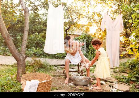 Charming kleines Mädchen helfen Mutter mit Weidenkorb während der Arbeit Chore und hängende Wäsche im Hinterhof im Sommer Abend Stockfoto