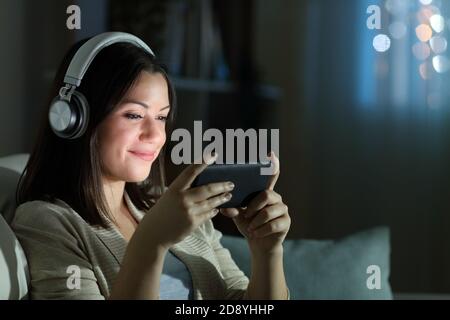 Entspannte Frau mit Kopfhörern, die Videos auf dem Smartphone ansieht Die Nacht auf einer Couch im Wohnzimmer sitzend Zu Hause Stockfoto