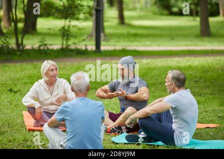 Die Gruppe der aktiven Senioren hat sich im Stadtpark versammelt Sitzen auf Matten und hören ihrem Yoga-Trainer zu Stockfoto