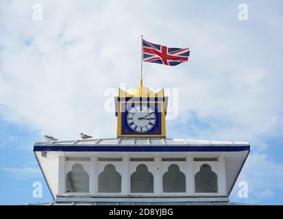 Die Union Jack Flagge fliegt auf dem Uhrenturm des Eastbourne Pier. Möwen sitzen bei Sonnenschein auf dem Dach. Stockfoto