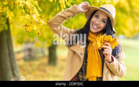 Glückliche dunkelhaarige Schönheit lächelt an einem Herbsttag mit gelben Blättern an der Kamera. Stockfoto