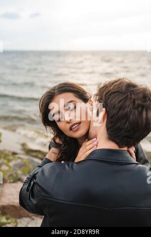 Mann in schwarzer Lederjacke küssen Freundin in Hals in der Nähe Meer am Strand Stockfoto