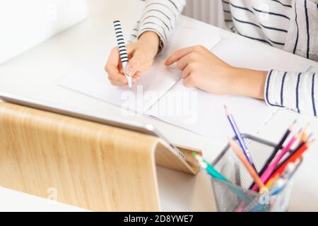 Nahaufnahme von Kinderhände, die Hausaufgaben im Notizbuch mit Stift schreiben. Kind sitzt am Tisch mit digitalem Tablet-Computer zu Hause Stockfoto