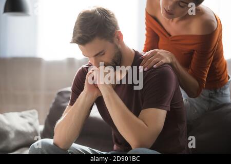 Junge Frau berührt Schulter des angespannten Mannes sitzend mit gebeugt Kopf und Hände in der Nähe des Gesichts halten Stockfoto