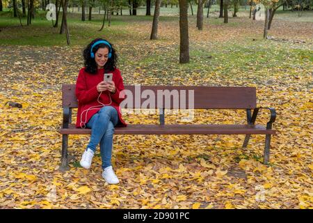 Junge Brünette Frau sitzt in einem Park Musik hören mit ihrem Handy und blauen Kopfhörern. Hintergrund von farbigen Bäumen und gefallenen Blättern Stockfoto