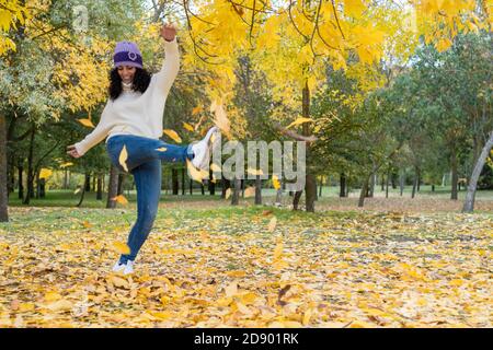 Junge Frau in einem Hut und weißen Wollpullover hat Macht Spaß, einen Haufen trockener Blätter zu treten, die gefallen sind Von den Bäumen in einem Park im Herbst Stockfoto