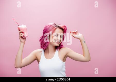Junge Frau mit bunten Haaren hält Erdbeermilchshake im Glas Mit Trinkhalm und Tanz isoliert auf rosa Stockfoto