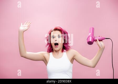 Verängstigte junge Frau mit bunten Haaren hält Fön isoliert auf Rosa Stockfoto