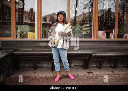 Modische afroamerikanische Frau in schwarzer Mütze und Fell mit zwei Baguette Rollen an den Händen. Stockfoto