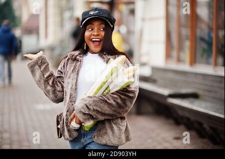 Modische afroamerikanische Frau in schwarzer Mütze und Fell mit zwei Baguette Rollen an den Händen. Stockfoto