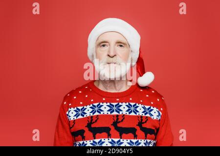 Senior Mann in santa Hut und Strickpullover Blick auf Kamera auf Rot isoliert Stockfoto