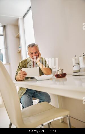 Smiley gealterter Gentleman, der während der Teezeit auf den Tablet-Bildschirm schaut Stockfoto
