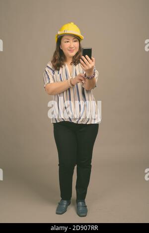 Reife schöne asiatische Geschäftsfrau trägt Hardhut vor grauem Hintergrund Stockfoto