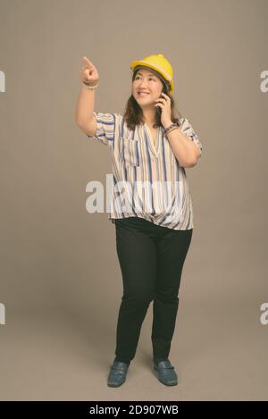 Reife schöne asiatische Geschäftsfrau trägt Hardhut vor grauem Hintergrund Stockfoto