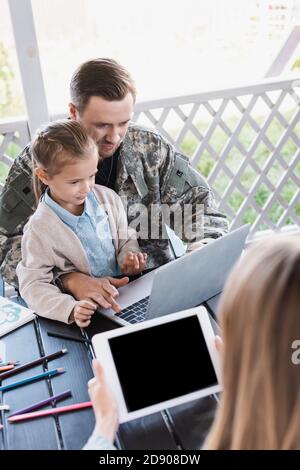 Militär Mann umarmt Mädchen und mit Laptop, während sitzen am Tisch mit verschwommenen Frau im Vordergrund Stockfoto