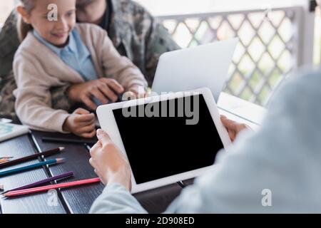 Frau hält digitales Tablet mit leerem Bildschirm mit verschwommenem Militär Mann und Tochter im Hintergrund Stockfoto