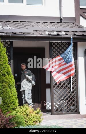 Ernsthafter Militärangehörige, der neben der Haustüre steht und wegschaut In der Nähe von Büschen und amerikanische Flagge Stockfoto