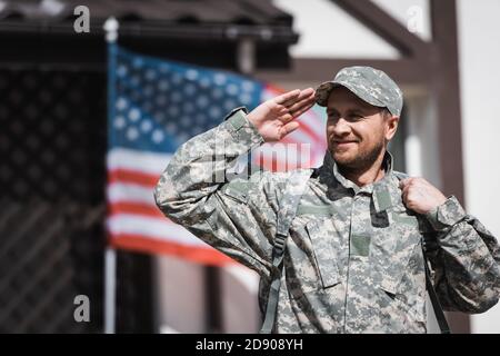 Lächelnder Militärmann in Uniform, der wegschaut und mit ihm grüsset Verschwommene flagge der usa auf dem Hintergrund Stockfoto