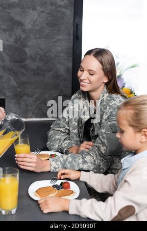 Mann Gießen Saft in der Nähe Frau in Militäruniform und Mädchen Sitzen am Tisch in der Küche Stockfoto