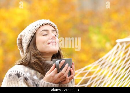 Entspannte Frau atmet frische Luft hält Kaffeetasse zum Frühstück Sitzen auf einer Seilhängematte im Herbst in einem Wald Stockfoto