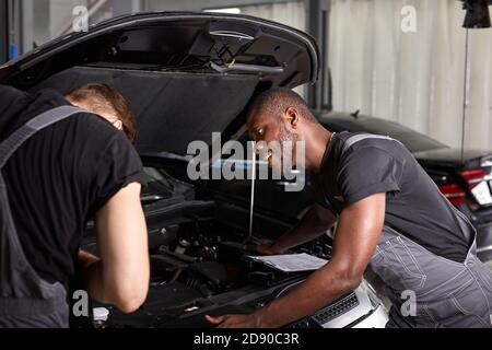 Zwei verschiedene Auto-Mechaniker, die beim Prüfen der Motorleistung in der Auto-Reparatur-Shop zusammenarbeiten, arbeiten zusammen, halten Diagnose Stockfoto