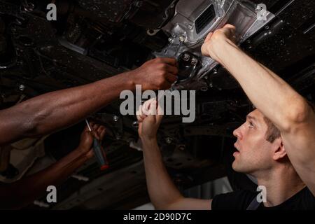 Blick von unten auf diverse Männer Reparatur Auto Boden, multi-ethnische Zusammenarbeit, zwei junge afrikanische und kaukasischen Jungs arbeiten zusammen. Stockfoto
