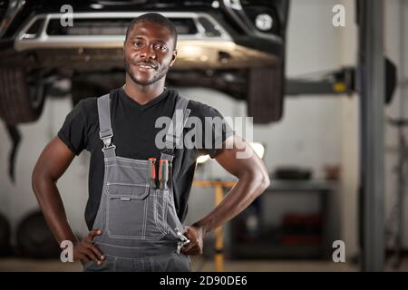 Professionelle schwarze Auto Mechaniker Blick auf Kamera, schöner schwarzer Kerl in Uniform ist scharf auf Reparatur Auto. Automobil im Hintergrund Stockfoto