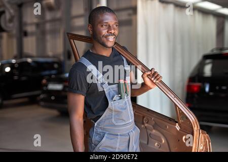 Junge afrikanische Automechaniker bei der Arbeit mit separaten Teil der Maschine, posiert bei der Kamera, gehen zu reparieren Stockfoto