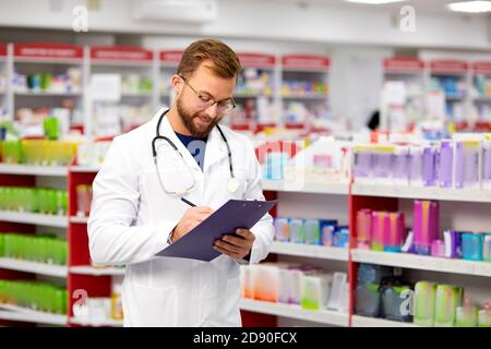 Angenehme männliche Apotheker Schreiben auf Zwischenablage bei der Apotheke, machen Sie Notizen, Porträt von schönen bärtigen Arbeiter der Apotheke in weißem Kleid Stockfoto