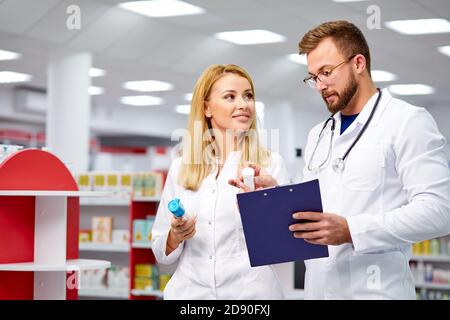 Zwei junge kaukasische Apotheker diskutieren Medikamente, mit einer digitalen Tablette und Dokumente. Mann und Frau in weißem medizinischen Kleid arbeiten in der Apotheke Stockfoto