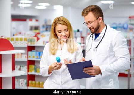 Zwei junge kaukasische Apotheker diskutieren Medikamente, mit einer digitalen Tablette und Dokumente. Mann und Frau in weißem medizinischen Kleid arbeiten in der Apotheke Stockfoto
