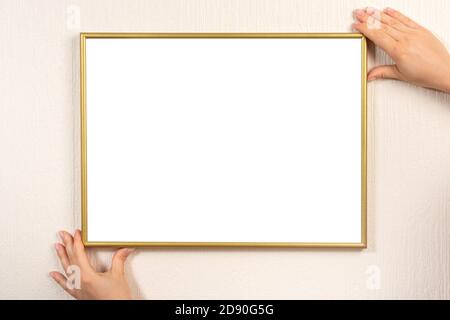 Goldener leerer Bilderrahmen, der an einer beigen Wand hängt. Hände hängen Fotorahmen mockup an der Wand. Junge Frau hält leeren Bilderrahmen mit Copyspace Stockfoto