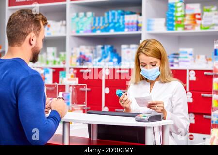 Junge schöne kaukasischen Apotheker in schützende medizinische Maske arbeitet mit Kunden in modernen Drogerie, dienen dem Kunden, trägt weiße medizinische Kleid Stockfoto