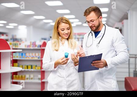 Zwei junge kaukasische Apotheker diskutieren Medikamente, mit einer digitalen Tablette und Dokumente. Mann und Frau in weißem medizinischen Kleid arbeiten in der Apotheke Stockfoto