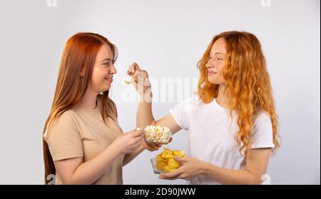 Genießen Sie Ihre Zeit. Rothaarige Freundinnen essen Chips und Popcorn, lachen und entspannen. Foto auf weißem Hintergrund. Stockfoto