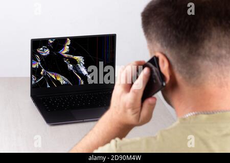 Ein junger Mann spricht auf einem Handy vor einem Laptop mit einem gebrochenen, rissigen Bildschirm in farbigen Flecken, das Konzept der Kontaktaufnahme mit technischen support Stockfoto