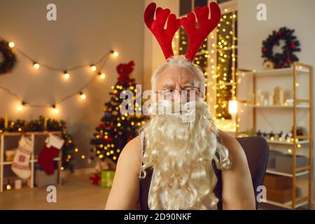 Portrait eines witzigen alten Weihnachtsmanns, der ein Hirschgeweih trägt und in einem gemütlichen Raum auf einem Stuhl sitzt. Stockfoto