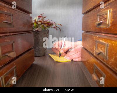 Nahaufnahme eines Mannes, der eine Notiz schreibt, betrachtet zwischen zwei alten hölzernen Schubladen auf einem Schreibtisch. Stockfoto