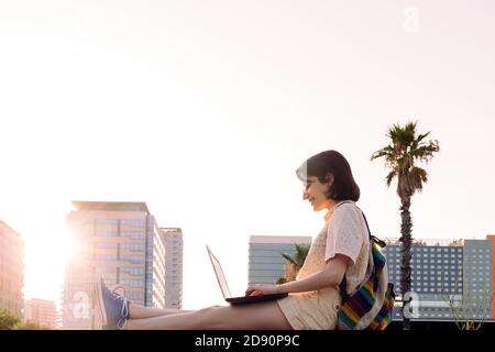 Glückliche junge Reisende Frau lächelt in der Stadt bei Sonnenuntergang mit ihrem Laptop und Rucksack, Konzept der Technologie, Jugend und digitalen Nomaden Lebensstil arbeiten Stockfoto
