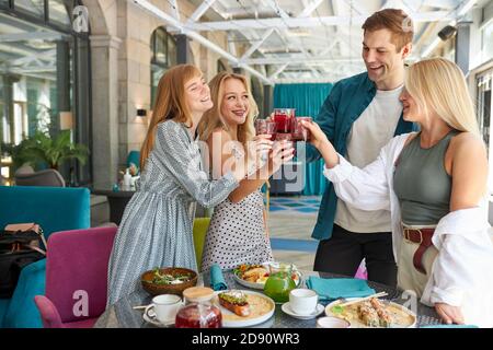 Gruppe von jungen Freunden klirren Gläser mit Getränken im Café, sie teilen Nachrichten, Ruhe nach der Arbeit oder feiern etwas Stockfoto