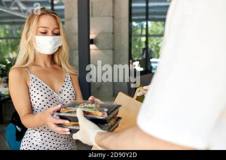 Liebenswert Frau Kunde nehmen ihre Bestellung im Restaurant während Coronavirus, Kellner des Cafés geben Essen in Behälter tragen Schutzhandschuhe, Frau ist in medizinischen Maske Stockfoto