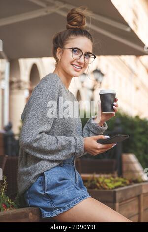 Junge lächelnde Frau in einer Brille mit Handy und Kaffeetasse zum Mitnehmen, die im Café im Freien steht. Stockfoto