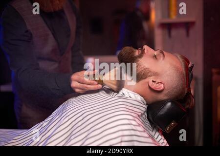 Junge kaukasische gut aussehende Mann auf dem Stuhl des Barbiers mit Verfahren der Rasur, erhalten Schönheitsverfahren in Friseur Salon Stockfoto